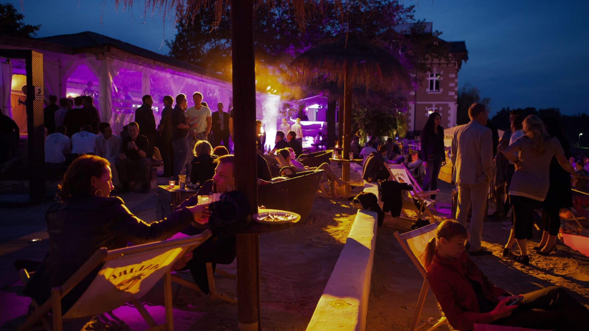 Outdoor evening nonprofit event with attendees enjoying drinks, socializing, and vibrant purple lighting, creating a lively atmosphere.