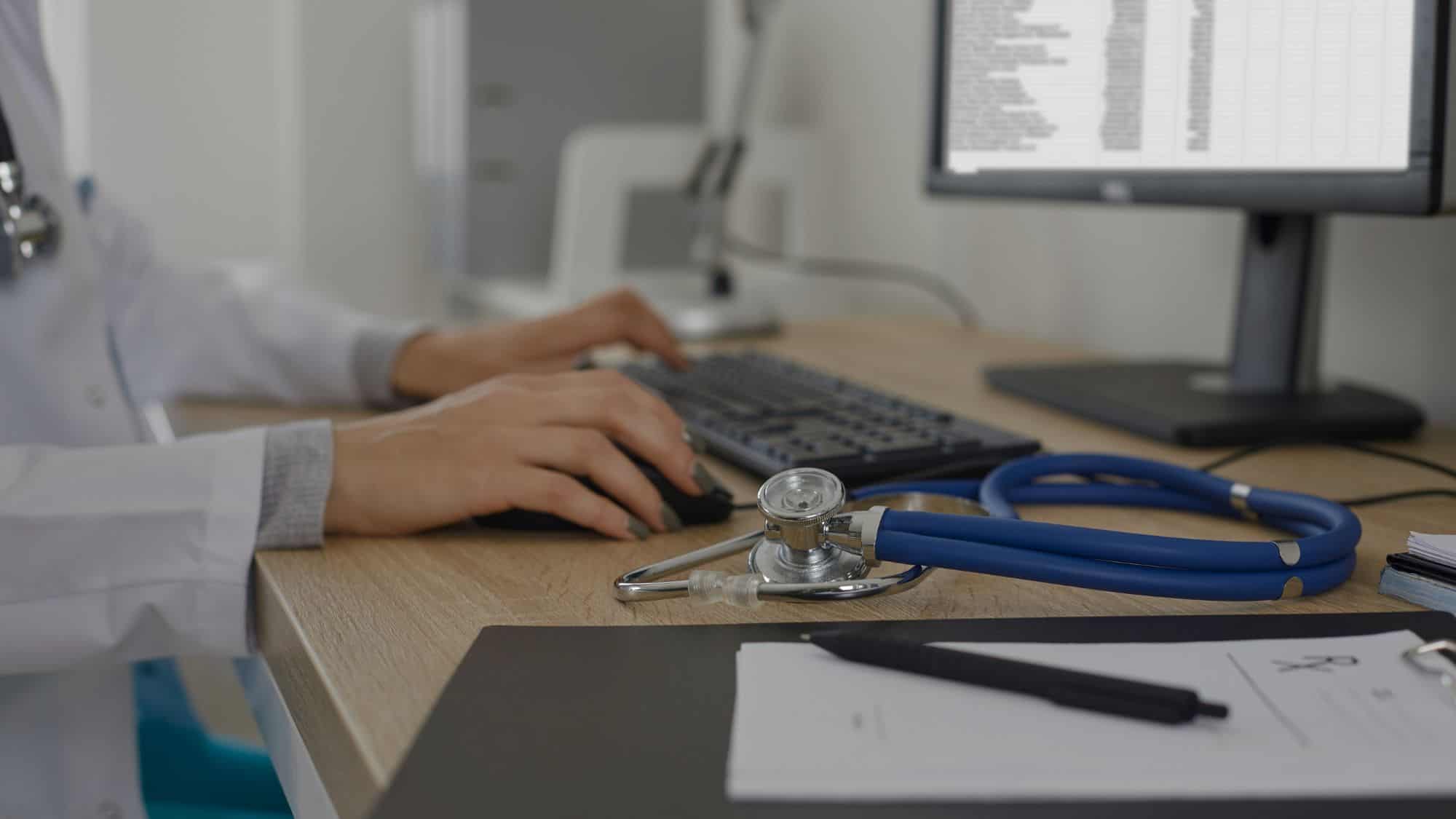 A doctor typing on a computer in a modern office setting, with a stethoscope and medical documents on the desk, representing the use of healthcare CRM software in the medical industry.