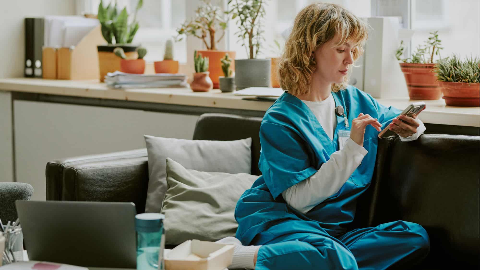 Healthcare professional in scrubs using a smartphone while sitting in a comfortable, plant-filled office space, symbolizing the integration of SMS marketing in healthcare.