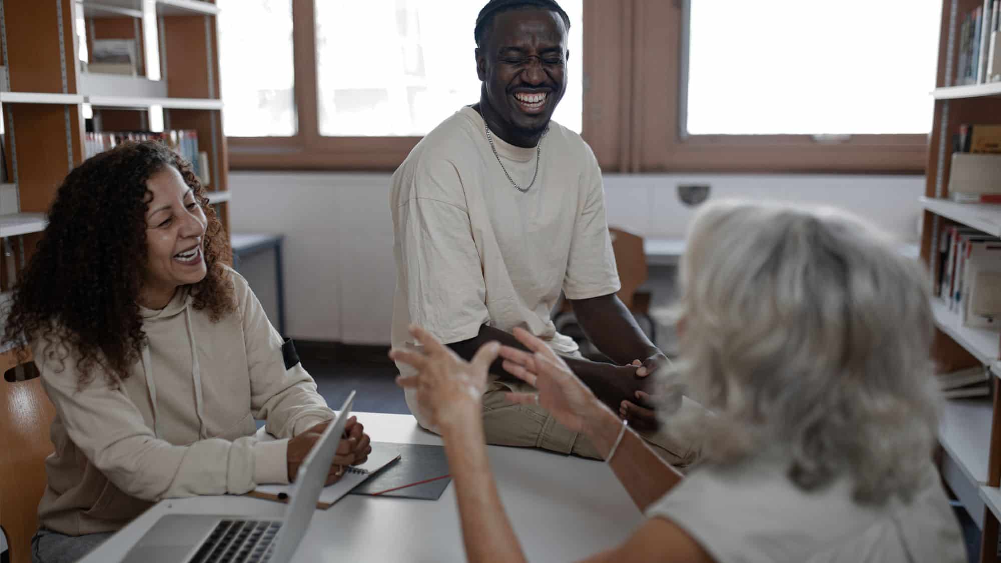 A group of three diverse individuals laughing and engaging in a discussion at a table in a library, symbolizing collaboration and brainstorming for digital marketing strategies tailored for nonprofits.
